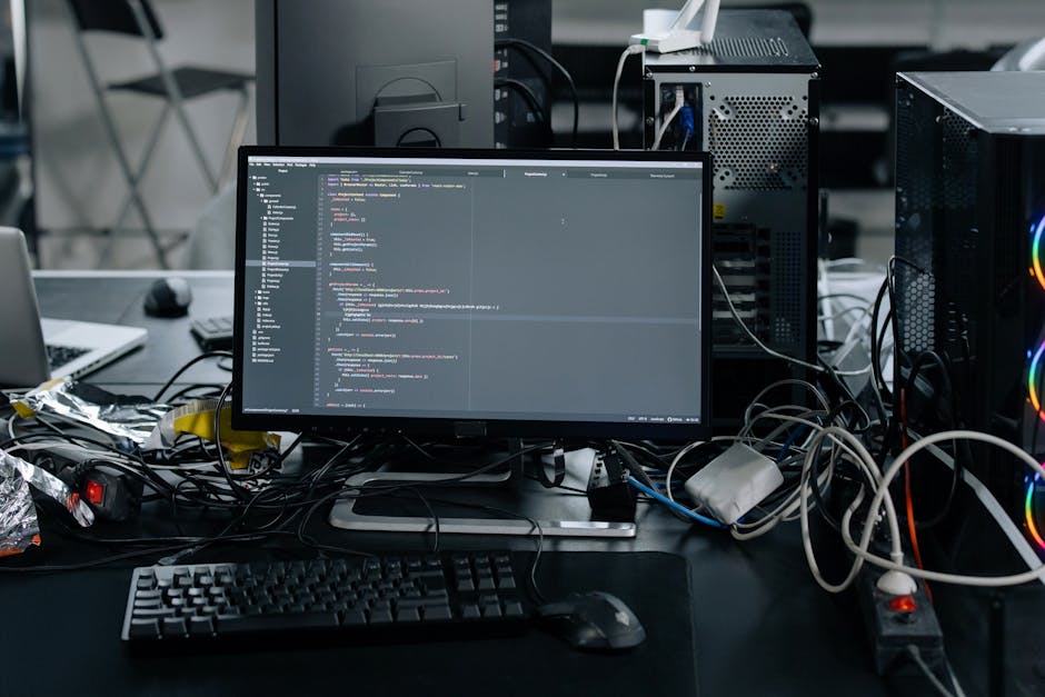 The image depicts a cluttered desk with a computer monitor displaying lines of code, surrounded by cables, a keyboard, and a mouse. To the right of the monitor are two computer towers with visible fan